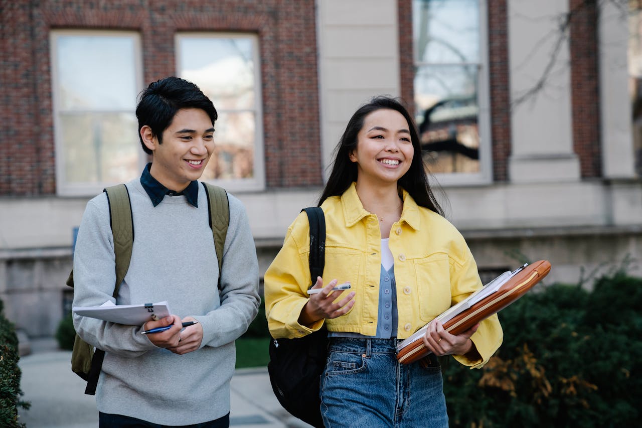 Two smiling students walking outdoors on a college campus, carrying supplies.